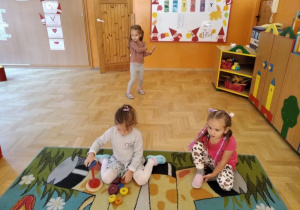 Dziewczynki siedzą na dywanie, w tle tańcząca dziewczynka.